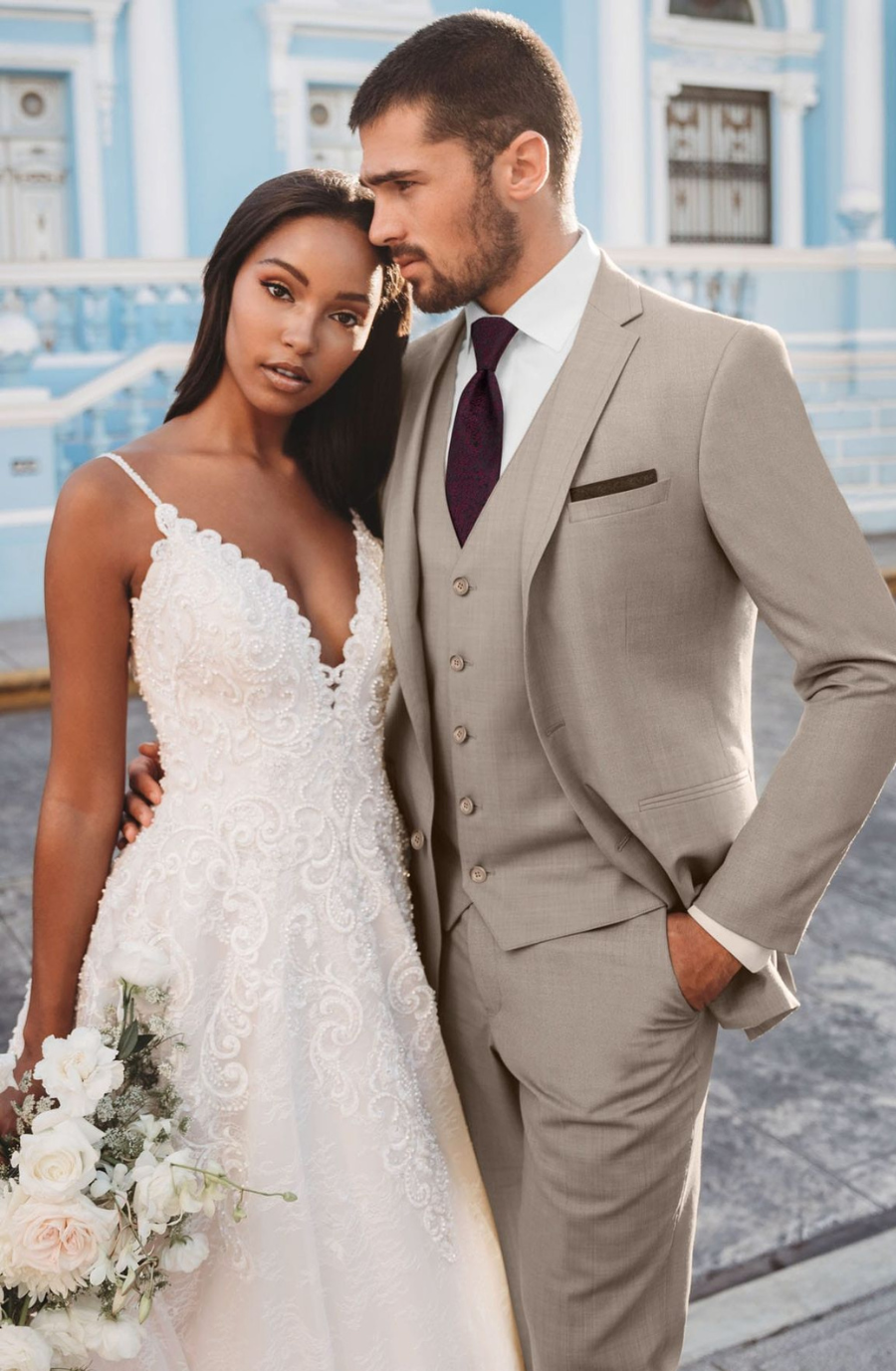 A female model wears a white lace spaghetti strap dress and male model wearings a light grey suit with a purple tie and white shirt