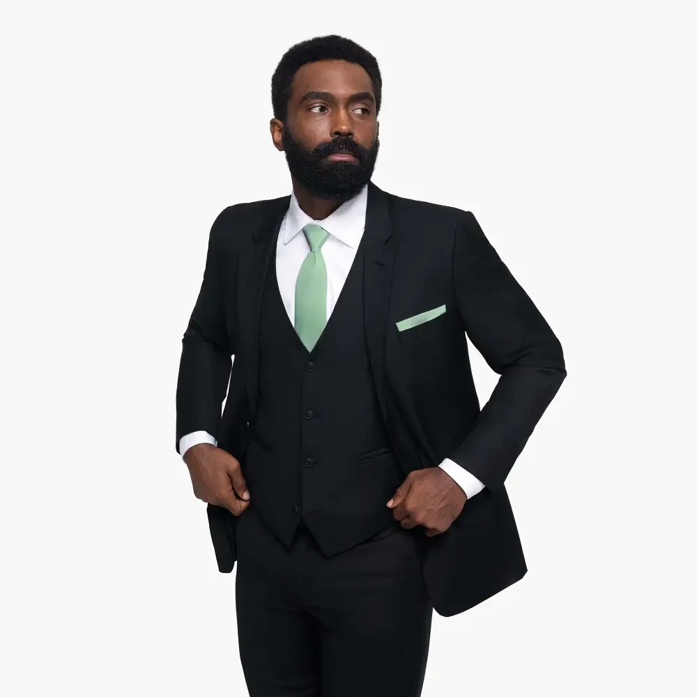 A man in a black suit with a green tie and pocket square, standing confidently with a slight smile.