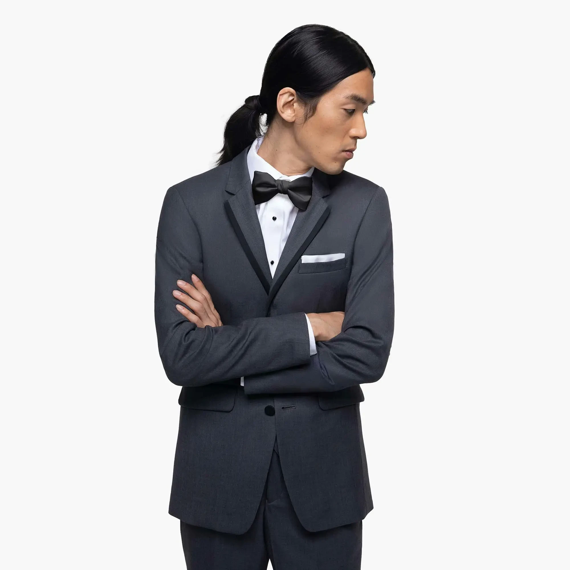 Man in a charcoal suit with a white shirt and black bow tie stands with arms crossed, facing left. He appears thoughtful, set against a plain background.