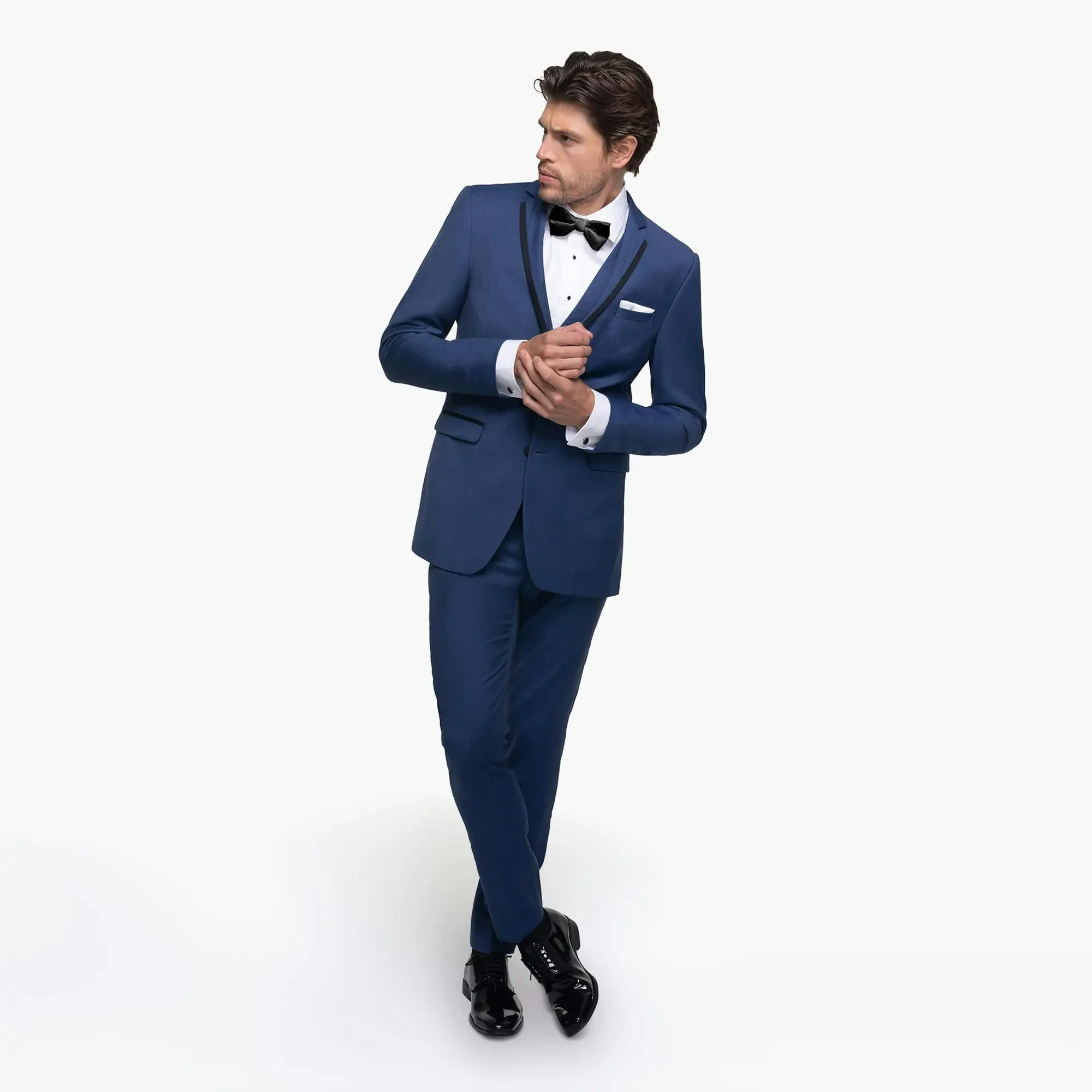 A man in a sleek blue suit with a black bow tie and white shirt poses confidently. His hands are clasped. The tone is stylish and formal.