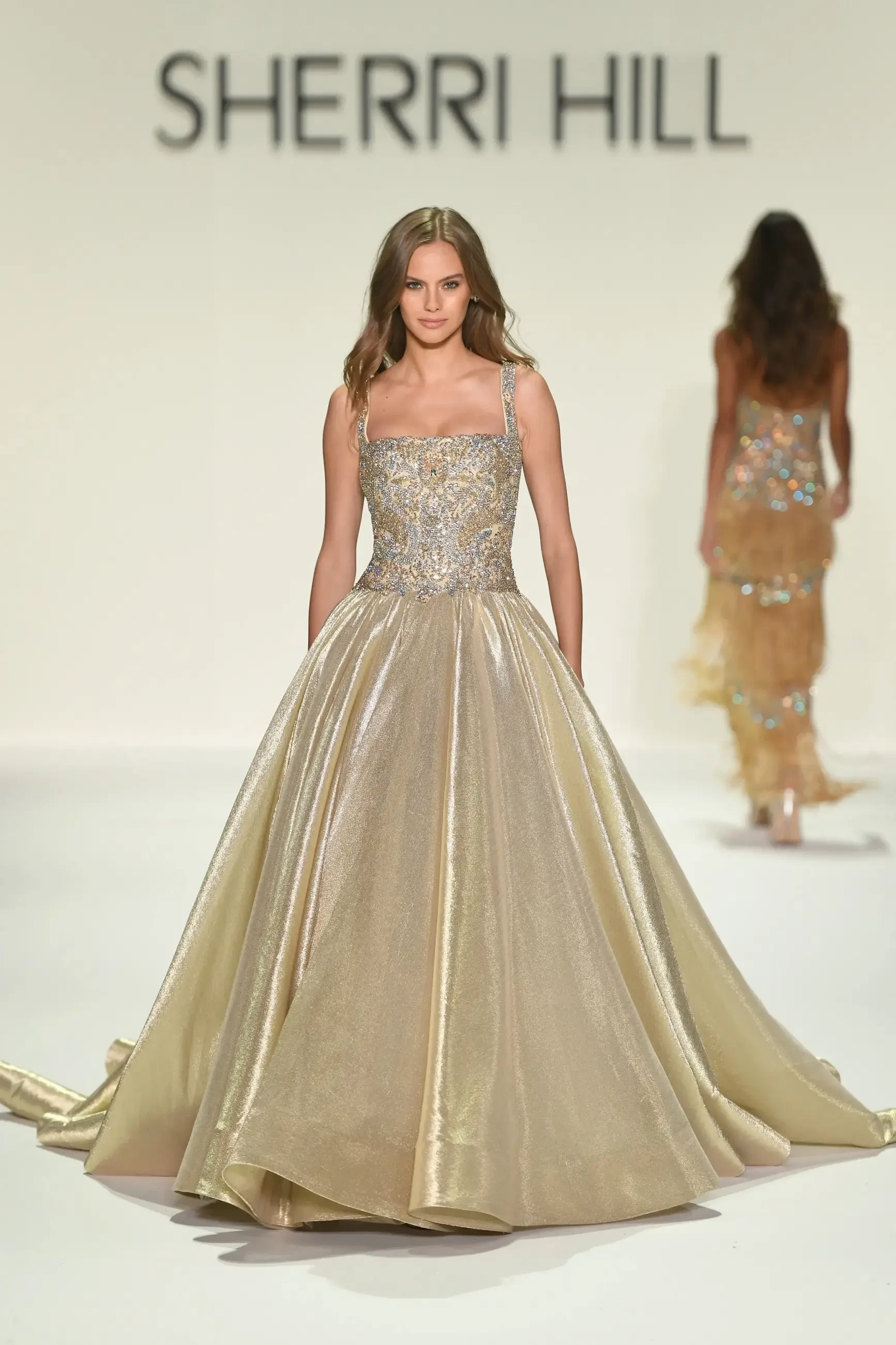 A model walks down the runway in a glamorous gown designed by Sherri Hill, featuring intricate detailing and a flowing skirt.
