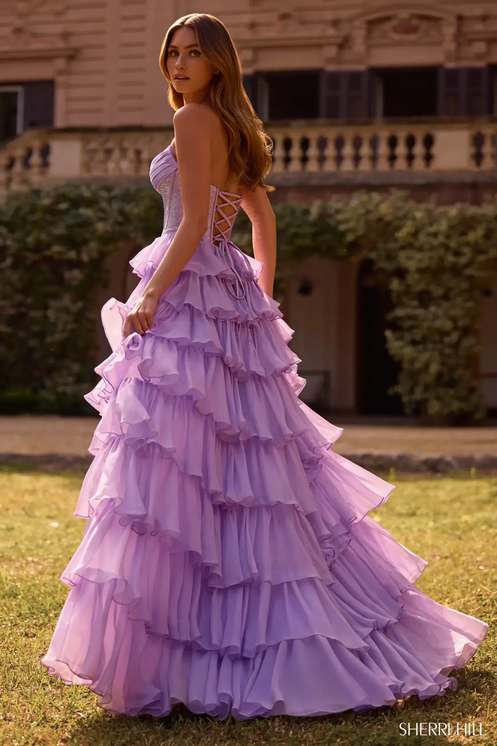A woman in a flowing lavender ruffled gown stands on grass, glancing back over her shoulder. Background features a stately building and greenery.
