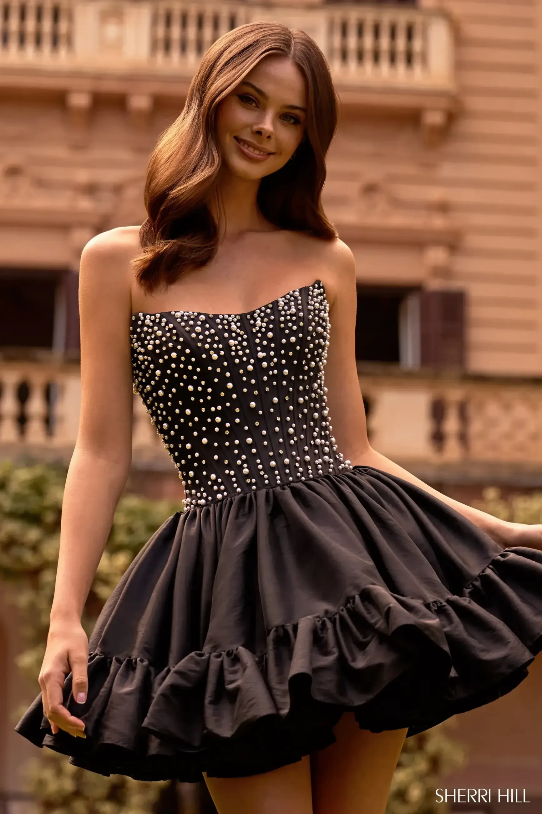 Model is wearing a black strapless cocktail dress with a pearl-embellished corset bodice and bubble skirt.