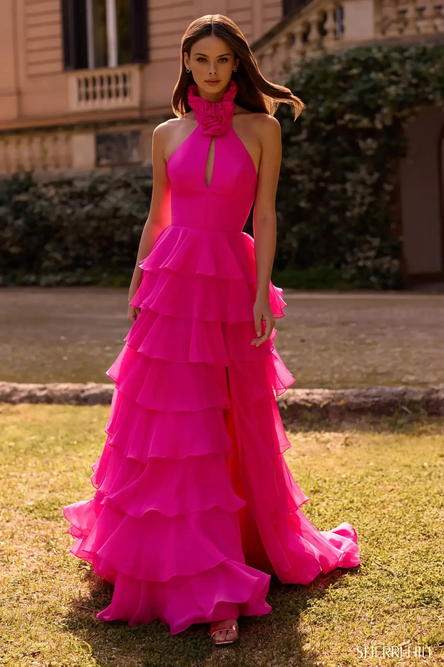 Model is wearing a vibrant halter chiffon gown with a keyhole cutout, rosette ruffle neckline, low open back, layered A-line skirt, and front slit.