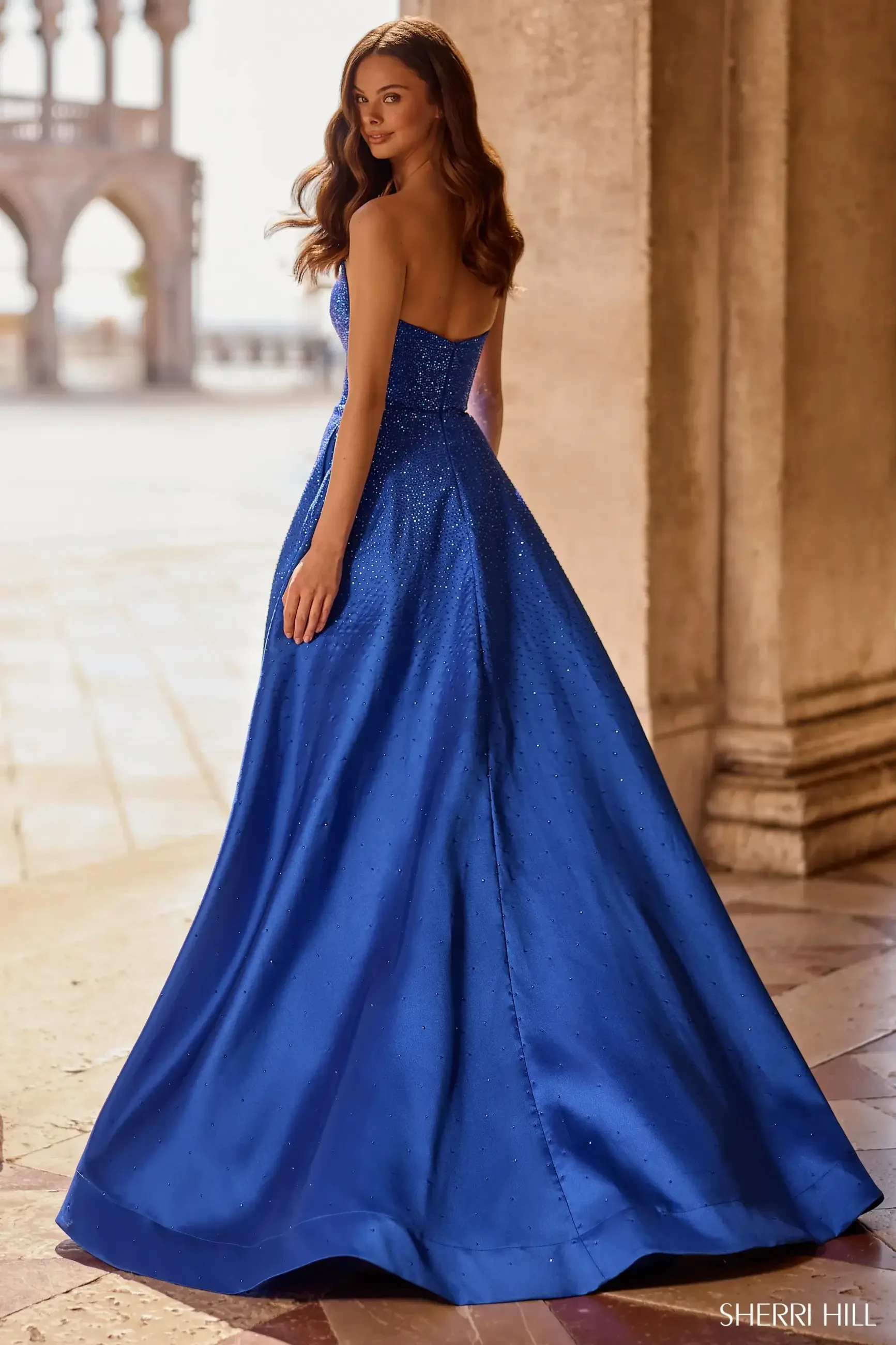 A woman in a blue gown turns away to show the back of the dress, standing in an elegant setting.