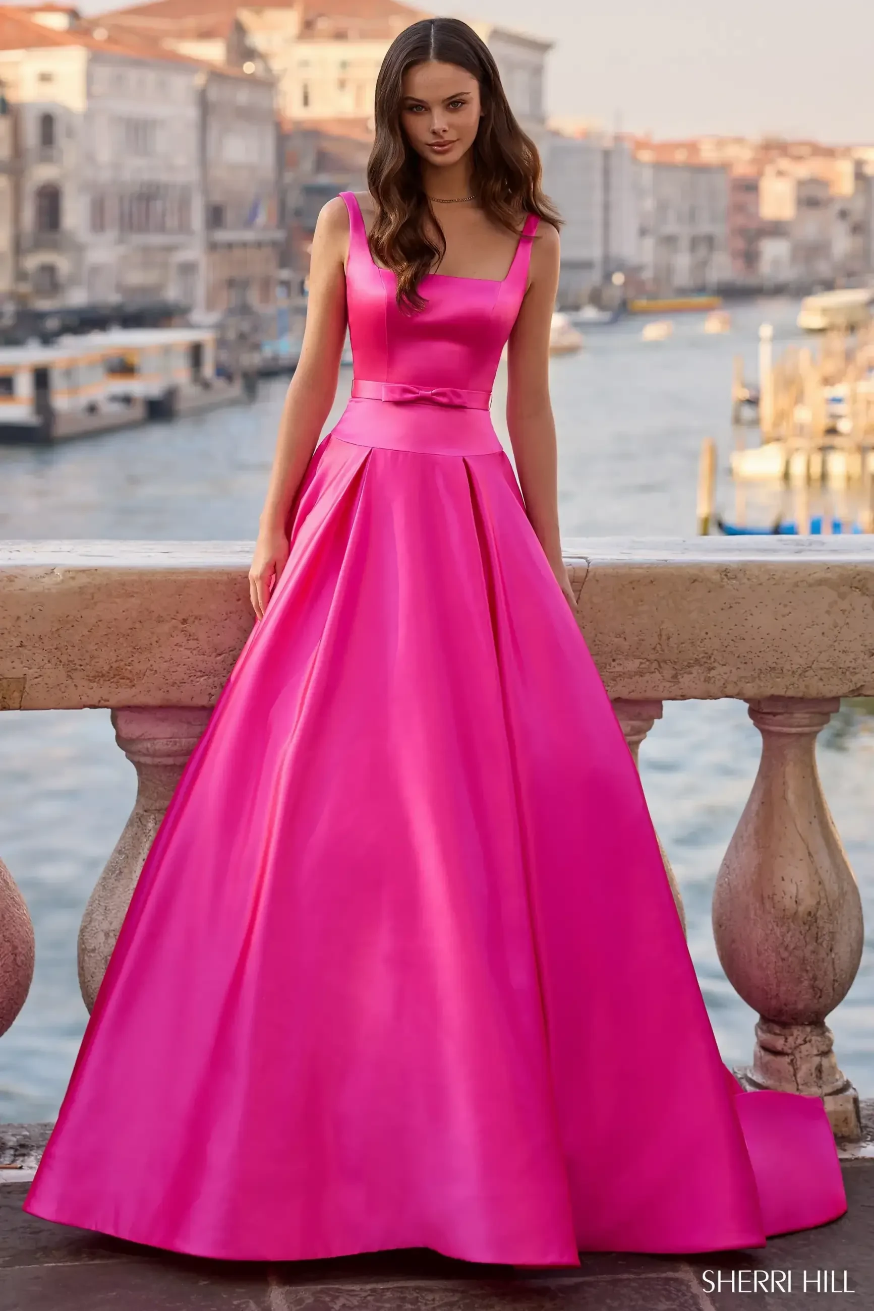 A young woman wearing a bright pink formal gown stands near a waterfront, with buildings and boats in the background.