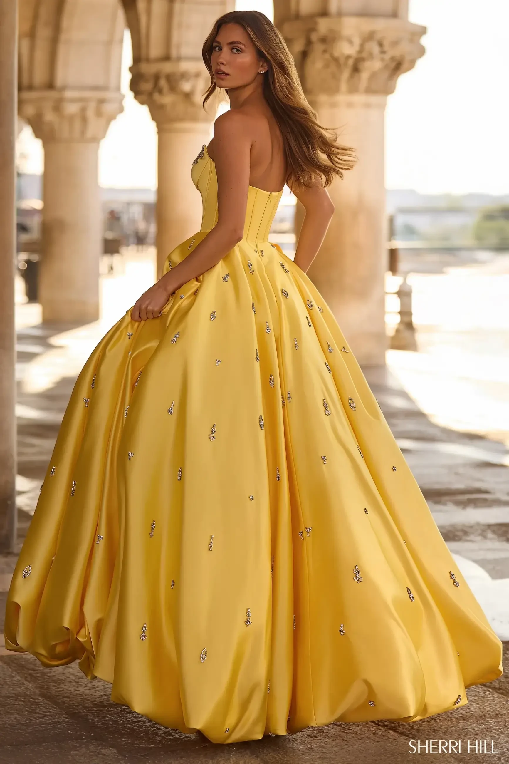 A woman in a strapless yellow ball gown stands gracefully in a sunlit stone corridor, exuding elegance and confidence. The dress is embellished with small sparkles.