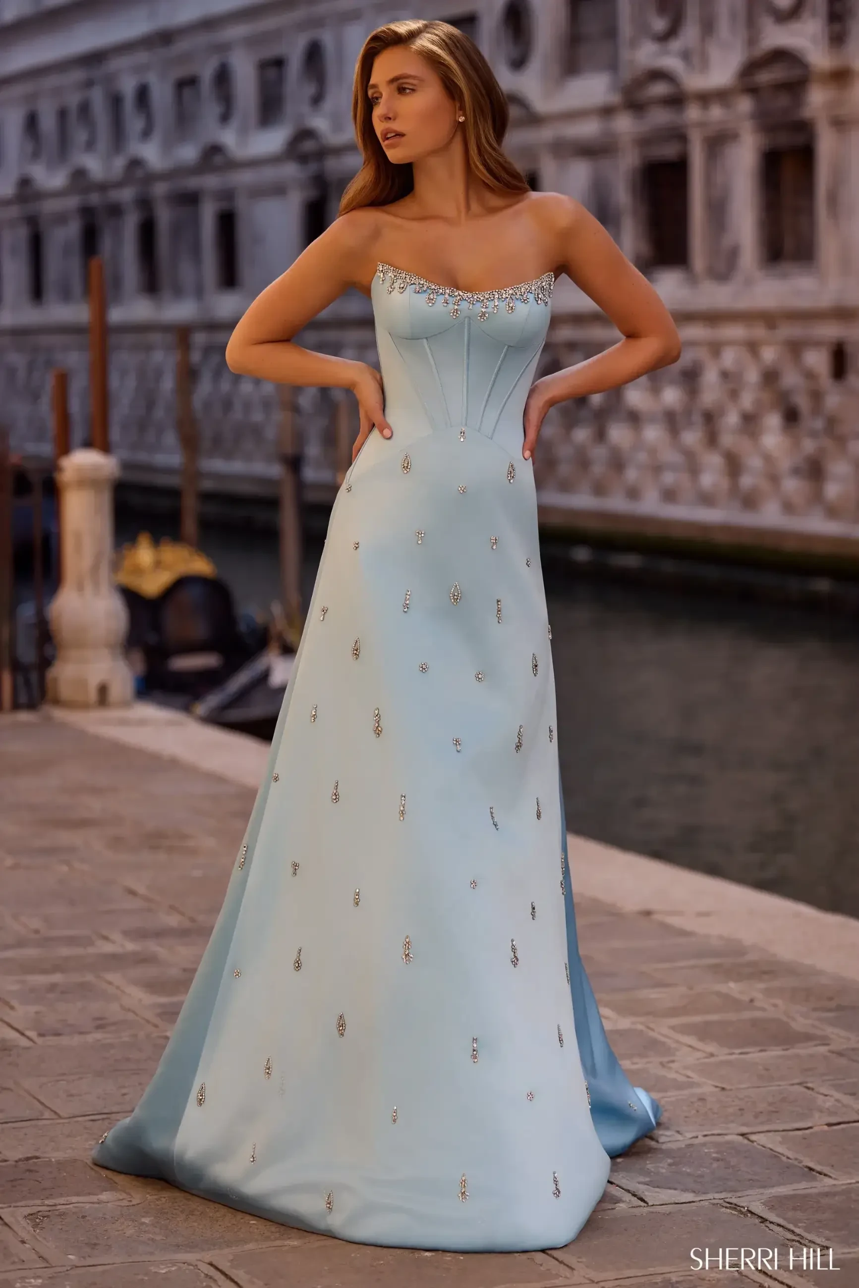 A woman in an elegant light blue gown with jeweled details stands on a cobblestone path near a canal. She looks off into the distance, exuding a serene and elegant aura.