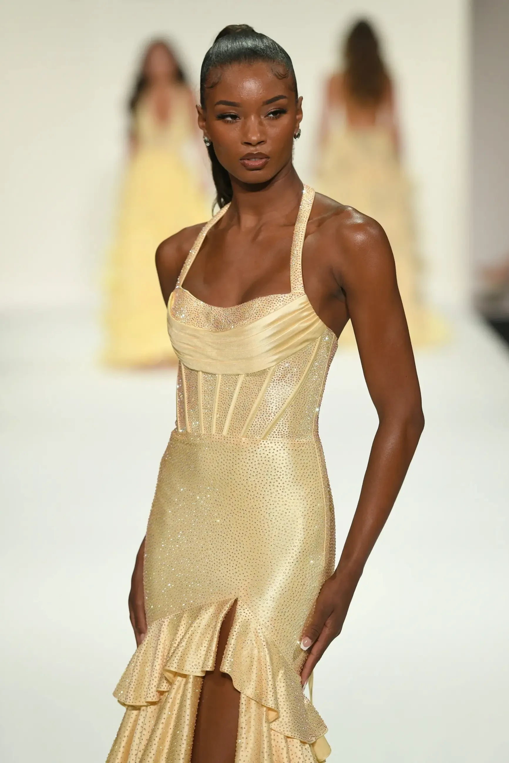 A model in a shimmering gold gown with a structured bodice and ruffled hem walks down a runway. The background shows blurred figures in similar attire.