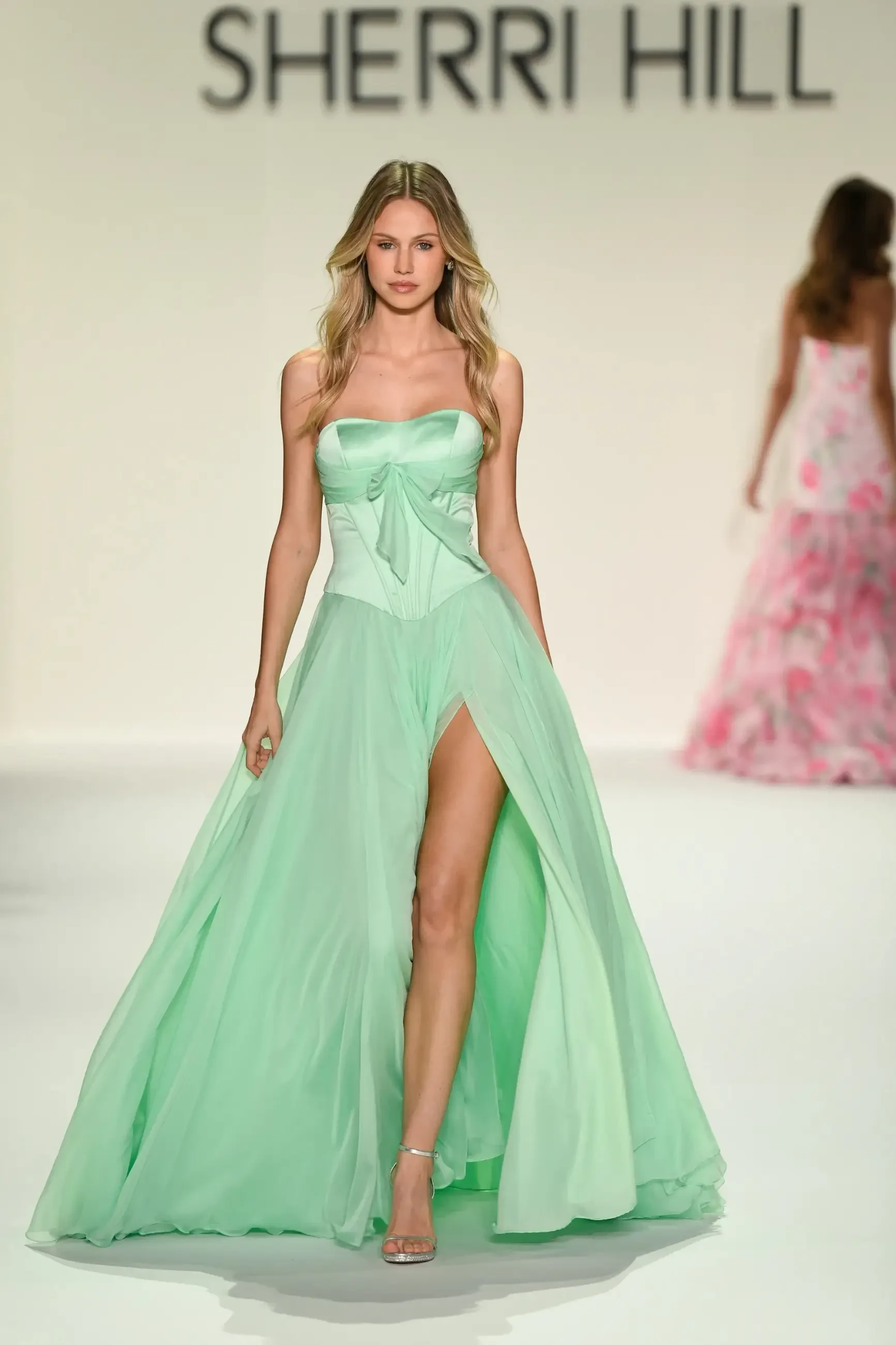 Female model walking on a runway in a flowing mint green dress with a high slit, under bright lighting. The background features blurred models in pastel gowns.