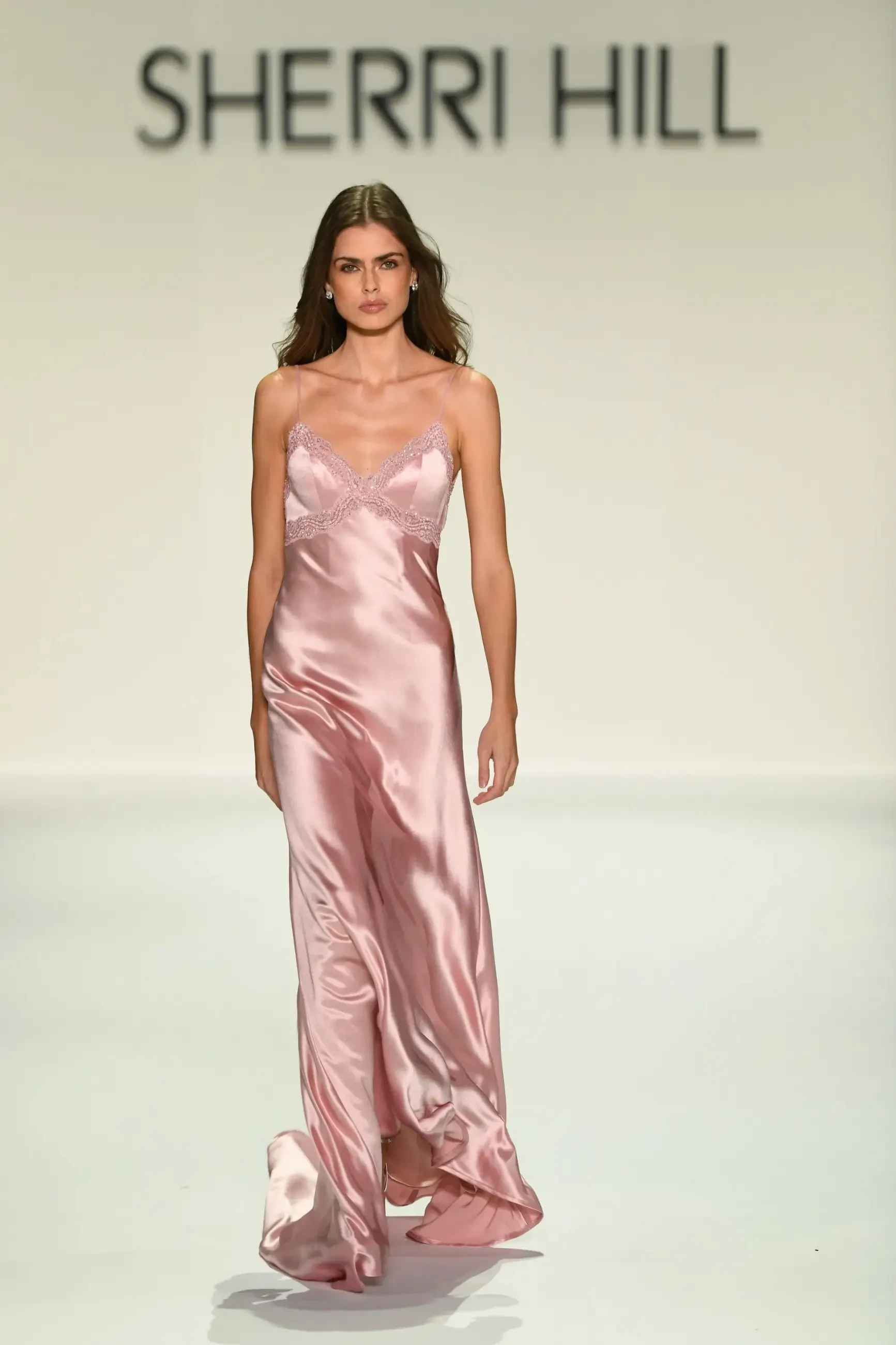 Model walks runway in a Sherri Hill pink satin gown with lace detail. The dress exudes elegance and confidence, set against a minimalistic backdrop.