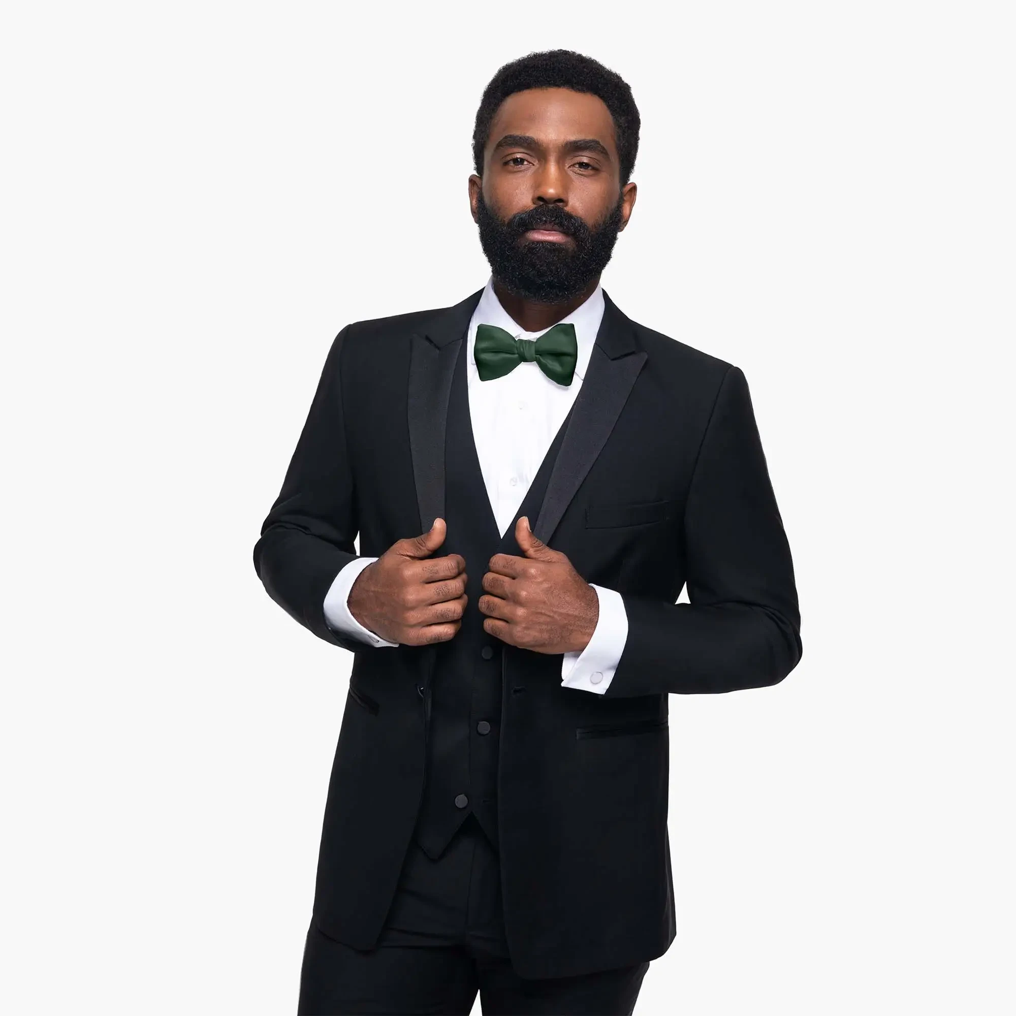 Man in a black tuxedo with a white shirt and green bow tie, adjusting his jacket. He has a full beard and a confident expression against a plain background.